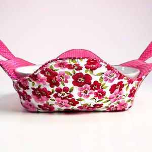 May include: A floral fabric bowl cozy with a white interior. The cozy features a white background with red and pink flowers and green leaves. It has pink handles with white polka dots. The cozy is designed to hold a bowl.