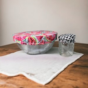 Op de afbeelding: Twee herbruikbare voedselhoezen op een houten tafel. Een hoes is een ronde, doorzichtige kom met een watermeloenprint stoffen deksel. De andere hoes is een glazen pot met een zwart-wit geruit stoffen deksel.