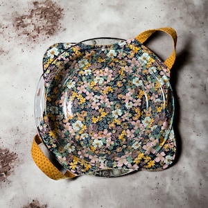 May include: A floral pie plate holder with yellow handles. The holder is made of fabric with a floral pattern and has a clear glass pie plate inside.