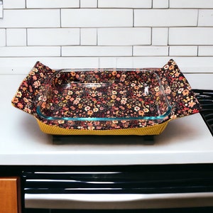 May include: A floral patterned fabric baking dish liner with a glass baking dish inside. The liner is yellow, brown, and black with a floral pattern. The baking dish is sitting on a white countertop.
