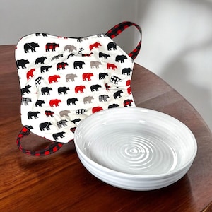 May include: A white ceramic bowl with a swirl pattern sits on a wooden table next to a red and black plaid fabric pot holder with a pattern of black bears.