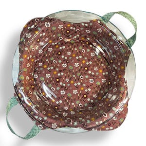 Puede incluir: Un molde para tarta de vidrio transparente con una cubierta de tela floral. La tela presenta un patrón de pequeñas flores blancas, amarillas y verdes sobre un fondo marrón. El plato tiene dos asas verdes con un estampado de puntos.