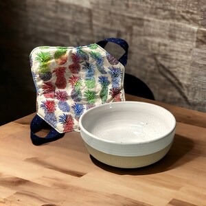 Może przedstawiać: Biała ceramiczna miska z kropkowanym szkliwem stoi na drewnianej powierzchni obok kolorowej rękawicy kuchennej z wzorem ananasa.