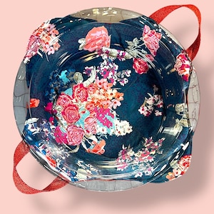 Puede incluir: Un cuenco de vidrio transparente con un forro de tela estampada floral azul oscuro. La tela presenta flores rosas, rojas y blancas. Las asas rojas brillantes están unidas al cuenco, creando una pieza decorativa y funcional.