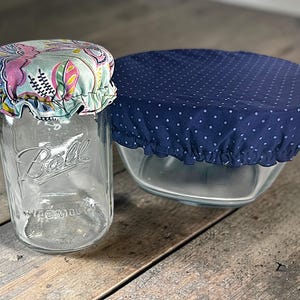 Op de afbeelding: Een heldere glazen Ball-pot met een bloemenstoffen deksel en een glazen kom bedekt met een marineblauwe polka dot stoffen deksel. De potdeksel heeft een lichtblauwe achtergrond met roze en gele bloemenmotieven. De komdeksel heeft een wit stippenpatroon.
