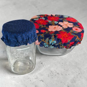 Op de afbeelding: Twee herbruikbare voedselhoezen. Eén is een marineblauwe stof met kleine blauwe stippen, die een glazen pot bedekt. De andere is een marineblauwe stof met een bloemenpatroon van rode, roze en blauwgroene bloemen, die een glazen kom bedekt.