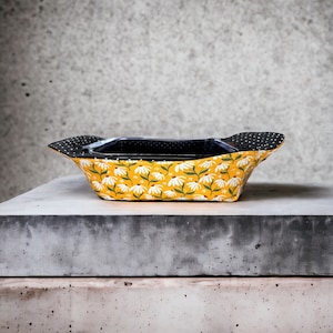 May include: A yellow ceramic baking dish with a white floral pattern and black rim. The dish is sitting on a grey concrete surface.