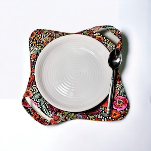 May include: A white ceramic plate with a spoon on a colorful floral fabric placemat. The placemat has a black background with red, yellow, and blue flowers.