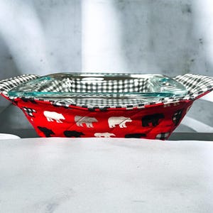 Puede incluir: Manopla para horno acolchada roja y negra con diseños de osos polares blancos. La manopla sostiene una fuente para hornear de vidrio transparente.