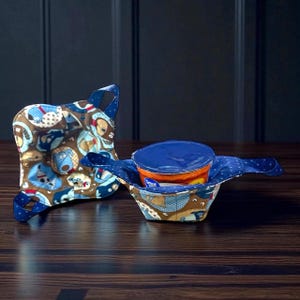 May include: Two fabric bowl cozies with a dog pattern. One is folded with a handle, the other is open and holding a blue-lidded container. The cozies are brown, blue, and white, with a repeating pattern of cartoon dogs.