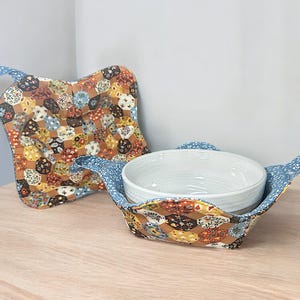 Puede incluir: Un salvamanteles acolchado y un agarrador a juego. El salvamanteles está diseñado para sostener un cuenco de cerámica blanco. Ambos artículos presentan un patrón de patchwork con diseños florales marrones, amarillos y azules. Los bordes están ribeteados con tela floral azul.