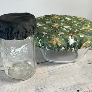 Op de afbeelding: Een heldere glazen pot met een zwarte stoffen deksel en een glazen kom bedekt met een groene stoffen deksel. De groene deksel heeft een patroon van kippen en andere boerderijdieren.