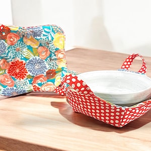 Puede incluir: Dos calientaplatos de tela. Uno es rojo con lunares blancos y el otro es turquesa con un estampado floral. Ambos tienen asas.