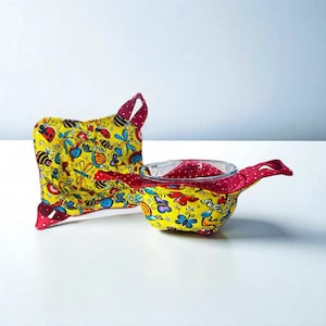 Può includere: Una presina gialla e un sottopentola quadrato abbinato. Entrambi sono decorati con un colorato motivo a insetti e farfalle. La presina ha dettagli e manici a pois rossi. L'interno del sottopentola è a pois rossi.