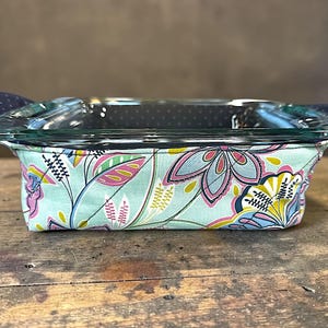 May include: A clear glass baking dish with a floral patterned fabric cover. The fabric features a light blue background with pink, yellow, and blue floral designs. The handles are navy blue with white polka dots. The dish is on a wooden surface.