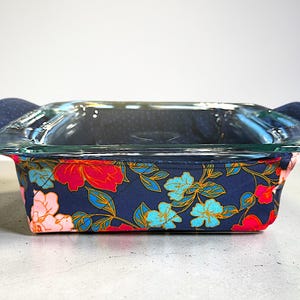May include: A clear glass baking dish with navy blue fabric floral print cozy. The fabric features red, pink, and turquoise flowers with green leaves. The dish has navy blue handles.