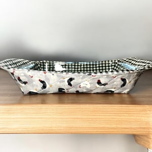 May include: A quilted fabric casserole dish with a black and white checkered lining and a rooster print. The dish is on a wooden surface.