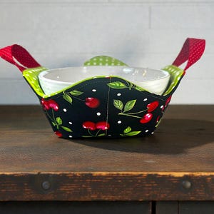 May include: A black bowl cozy with a cherry pattern, white polka dots, and green leaves. The cozy has red handles with white polka dots and holds a white bowl. The interior is green with white polka dots.