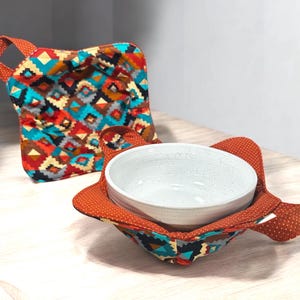 May include: Two colorful fabric pot holders with geometric patterns. One pot holder is folded and the other is holding a white ceramic bowl.