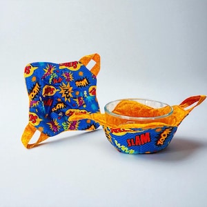 May include: Two blue and yellow fabric bowl cozies with comic book-style graphics. One is square, the other is wrapped around a clear glass bowl. The fabric has words like "Zoom" and "Slam".