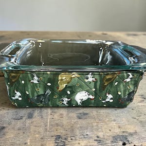 May include: A clear glass baking dish with dark gray handles, resting in a green quilted fabric cozy. The fabric features a pattern of chickens, white dots, and olive-green leaves. The dish sits on a wooden surface.