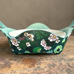 Puede incluir: Un protector de cuenco con estampado floral y asas color turquesa. El protector presenta un fondo verde oscuro con flores blancas, moradas y amarillas. Las asas y el interior tienen un estampado de cuadros turquesa y blanco. Dentro hay un cuenco blanco.