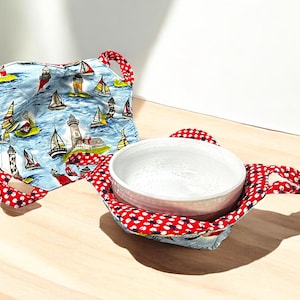 Puede incluir: Un calientaplatos de tela rojo y blanco con un forro de tela azul y blanco con un tema náutico con faros y veleros. El calientaplatos contiene un cuenco de cerámica blanco.