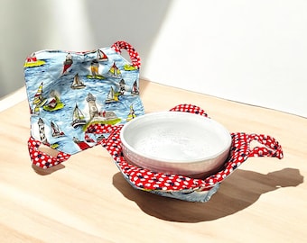 May include: A red and white fabric bowl cozy with a blue and white fabric liner featuring a nautical theme with lighthouses and sailboats. The cozy is holding a white ceramic bowl.