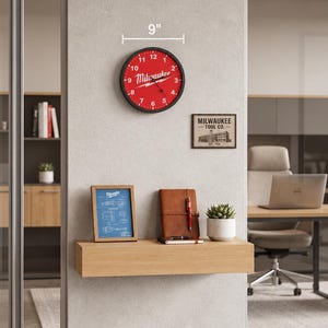May include: A round wall clock with a black frame and a red face featuring the white Milwaukee logo and numbers 1 through 12. The clock has a diameter of 9 inches. Below it, a rectangular wooden shelf holds a blue framed print, a brown notebook with a pen, and a small potted succulent plant.