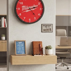 May include: A round wall clock with a red face, black frame, and white numbers featuring the Milwaukee logo and a lightning bolt graphic. Below it, a small framed print labeled Milwaukee Tool Co., a blue blueprint, a brown notebook, and a succulent.