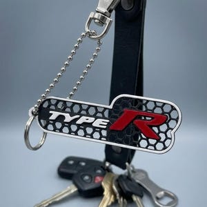 May include: A black leather keychain strap with a silver metal lobster clasp and a silver ball chain. Attached is a rectangular metal tag featuring a black honeycomb pattern, the white text "TYPE", and a large, bold, red letter "R" emblem.