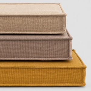 May include: A stack of three rectangular floor cushions featuring a textured fabric finish. The top cushion is light beige, the middle cushion is a muted mauve, and the bottom cushion is a vibrant mustard yellow. Each cushion has piped edges.