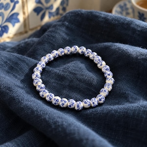 Puede incluir: Una pulsera circular compuesta por pequeñas cuentas esféricas blancas, cada una con un delicado patrón floral azul. Las cuentas están ensartadas en un bucle continuo, creando un diseño uniforme que resalta el contraste entre la base blanca y los intrincados motivos botánicos azules impresos en cada cuenta individual.