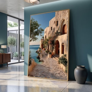 May include: A large rectangular wall art print in a thin gold frame depicting a Mediterranean coastal scene with stone buildings, a cobblestone path, and terracotta pots along a blue sea under a clear sky, displayed in a modern room.