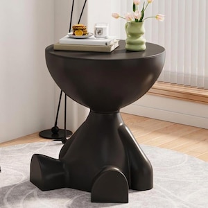 May include: A matte black side table shaped like a seated figure with a round tabletop. On the table sits a stack of two books, a small plate with orange slices, a clear glass candle holder, and a light green vase containing pink tulips.
