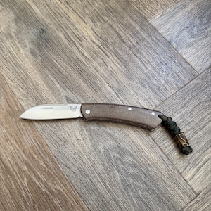 Puede incluir: Una navaja plegable con una hoja plateada que muestra el logotipo de Benchmade, un mango texturizado de color marrón oscuro asegurado con tres remaches plateados y un cordón de paracord verde oscuro con una cuenta metálica.