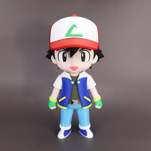 May include: A stylized vinyl figure of a character with spiky black hair, wearing a red and white cap with a green L-shaped emblem, a blue vest over a white shirt, light blue pants, and green fingerless gloves, standing in a neutral pose.
