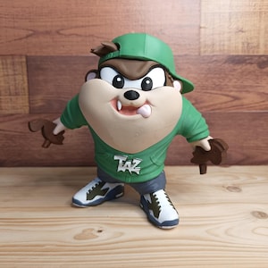 May include: A figurine of the Tasmanian Devil character wearing a green baseball cap, a green hooded sweatshirt with the text TAZ printed on the front, dark gray shorts, and white and green sneakers. The character has brown fur and white teeth.