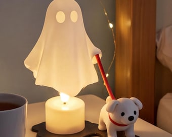 Op de afbeelding: Een witte spookvormige lamp met twee ovale ogen op een zwarte voet naast een klein wit hondenfiguurtje met een rode halsband. Het spook houdt een rode riem vast die aan de hond is bevestigd en de lamp wordt verlicht door een lichtbron.