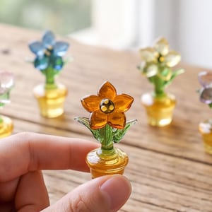 May include: A miniature glass flower figurine with five orange petals and a dark center, featuring a green stem and a yellow pot. Four additional glass flower figurines in blue, yellow, purple, and pink are arranged in the background.