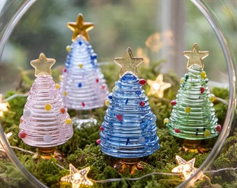 Op de afbeelding: Vier miniatuur kerstbomen van glas in roze, lichtblauw, donkerblauw en groen, elk met een spiraalontwerp, kleurrijke glazen stippen en een gouden ster als piek. Deze ornamenten staan in een glazen terrarium gevuld met mos.