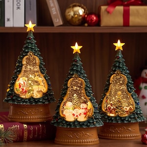 Könnte beinhalten: Drei konische Weihnachtsbaumdekorationen in Dunkelgrün mit strukturierter Nadeloptik. Jeder Baum verfügt über ein beleuchtetes Sichtfenster mit festlichen Szenen, einen goldenen Sockel mit Verzierungen und einen leuchtenden gelben Stern.