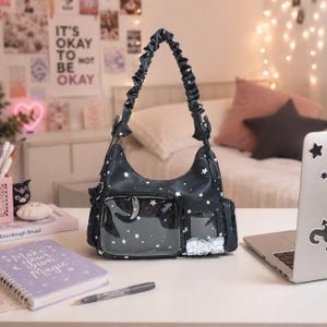 May include: A black shoulder bag featuring a white star pattern, a gathered scrunchie-style strap, and two clear vinyl front pockets. One pocket is rectangular and the other is smaller with a white ruffled trim detail along the bottom edge.