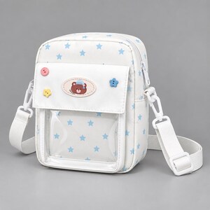 May include: A white crossbody bag featuring a light blue star pattern, a clear front pocket, and a flap closure. The flap is decorated with a small oval patch showing a brown bear wearing a blue nightcap, along with pink, yellow, and blue star-shaped buttons.