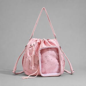 May include: A light pink drawstring backpack featuring a star-patterned fabric, a clear rectangular front pocket, and a star-shaped charm hanging from a side strap. The bag includes adjustable shoulder straps and silver-toned metal hardware.