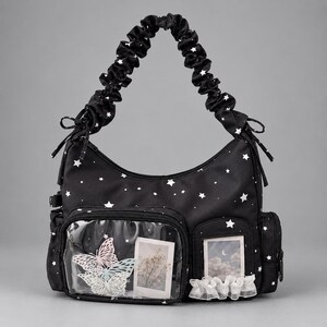 May include: A black shoulder bag featuring a white star pattern and a gathered, scrunchie-style strap. The front includes two clear display pockets, one containing butterfly cutouts and a photo, and the other holding a photo with white ruffles.
