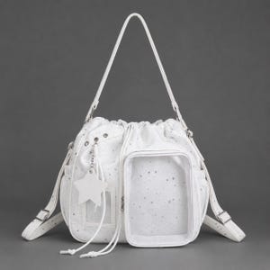 May include: A white drawstring backpack featuring a silver star pattern, a clear rectangular front pocket, and a star-shaped charm hanging from a silver chain. The bag includes adjustable shoulder straps with silver metal buckles and hardware.