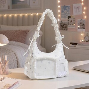 May include: A white hobo bag featuring a subtle grey star pattern, a gathered scrunchie-style shoulder strap with side ties, a clear rectangular front pocket, and a smaller side pocket with a white ruffled trim detail on the front exterior.