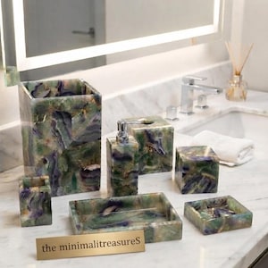Può includere: Un set di accessori da bagno realizzato in pietra verde e viola con venature naturali, che include un cestino alto, un dispenser di sapone con pompa argentata, un copriscatola per fazzoletti, un portaspazzolino, un portasapone e un piccolo vassoio quadrato.
