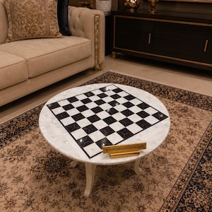 Puede incluir: Una mesa de centro redonda con una superficie de nácar blanco y un patrón de tablero de ajedrez en blanco y negro en el centro. La mesa tiene patas blancas curvas y una pequeña placa dorada con el texto THEMINIMALITREASURES.
